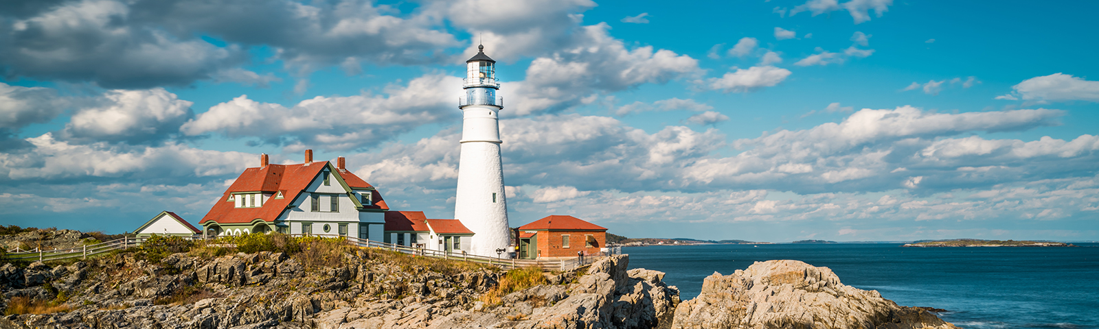 A coastal scene featuring a red and white lighthouse with a white building, surrounded by the ocean under a partly cloudy sky.