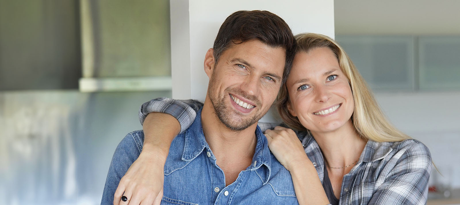 A man and a woman are embracing each other, with the man in the foreground and the woman behind him. They appear to be in a home setting, as suggested by the kitchen countertop visible in the background. The image has an overlaid watermark text that reads  alt  across it.