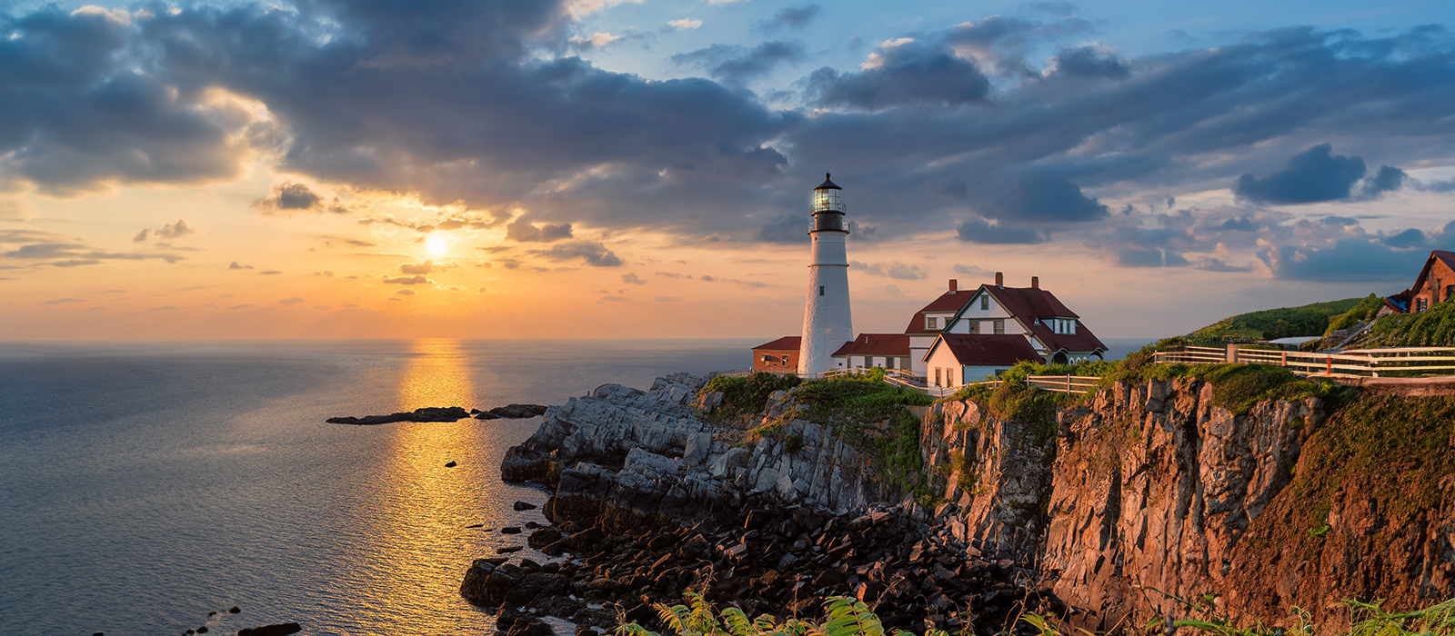 Lighthouse overlooking vast ocean at sunset, with a clear sky and lush greenery.