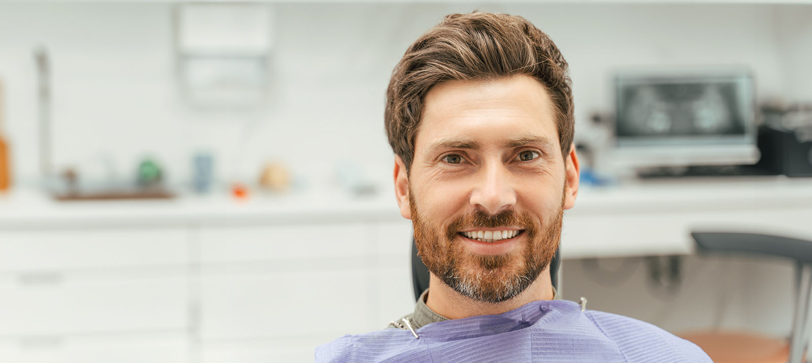 A man in a dental chair, smiling and wearing a red beard.
