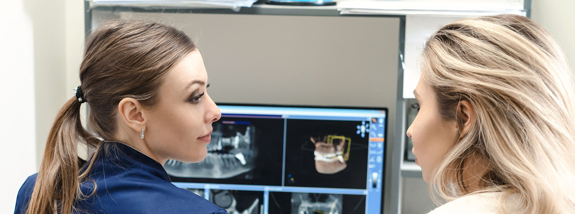 Two women in a medical setting, one wearing a blue shirt and the other with blonde hair, looking at an X-ray machine while standing next to each other.