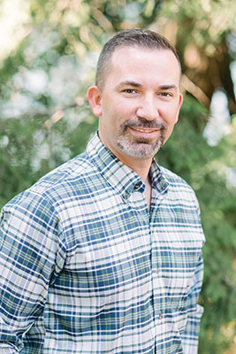 The image shows a man with a beard, wearing a plaid shirt and a blue lanyard around his neck, standing outdoors in front of trees.