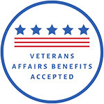 The image features a circular emblem with a blue background, containing a design resembling a shield with red and white stripes at the bottom. Inside the shield, there is text that reads  VETERANS AFFAIRS SUPPORTIVE SERVICES  in capital letters, followed by  APPROVED  in smaller font size below it. The emblem has a border with stars around its perimeter and is adorned with a red ribbon at the bottom.