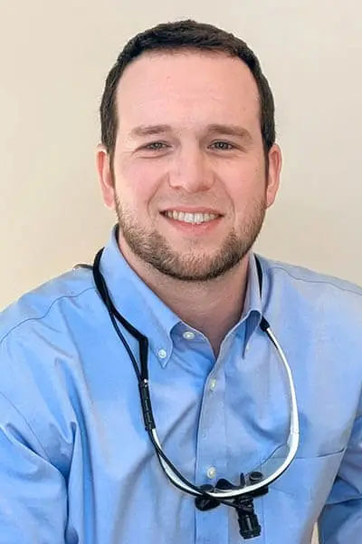 The image is a photograph of a man smiling at the camera. He has short, dark hair and is wearing a light blue shirt with a collar, a white lanyard with an identification badge, a pair of sunglasses hanging from his neckline, and a strap of what appears to be a water bottle around his waist. The background is neutral and out of focus, emphasizing the subject in the foreground.