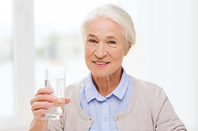 The image shows an elderly woman holding a clear glass of water, smiling and looking directly at the camera.