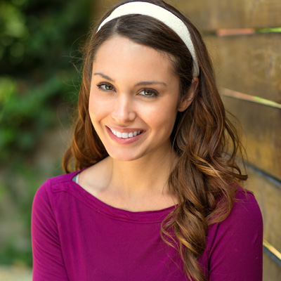 A smiling woman with long hair, wearing a purple top and headband, posing against a wooden fence.