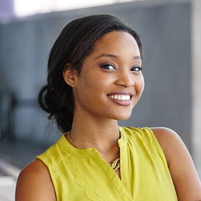 A smiling woman with dark hair, wearing a yellow top and a necklace, poses against a building backdrop.