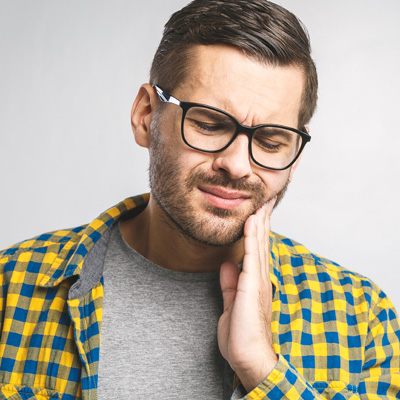 The image shows a man with glasses, a beard, and a mustache holding his hand to his mouth. He is wearing a yellow plaid shirt and appears to be in pain or discomfort, as suggested by his facial expression and hand placement.