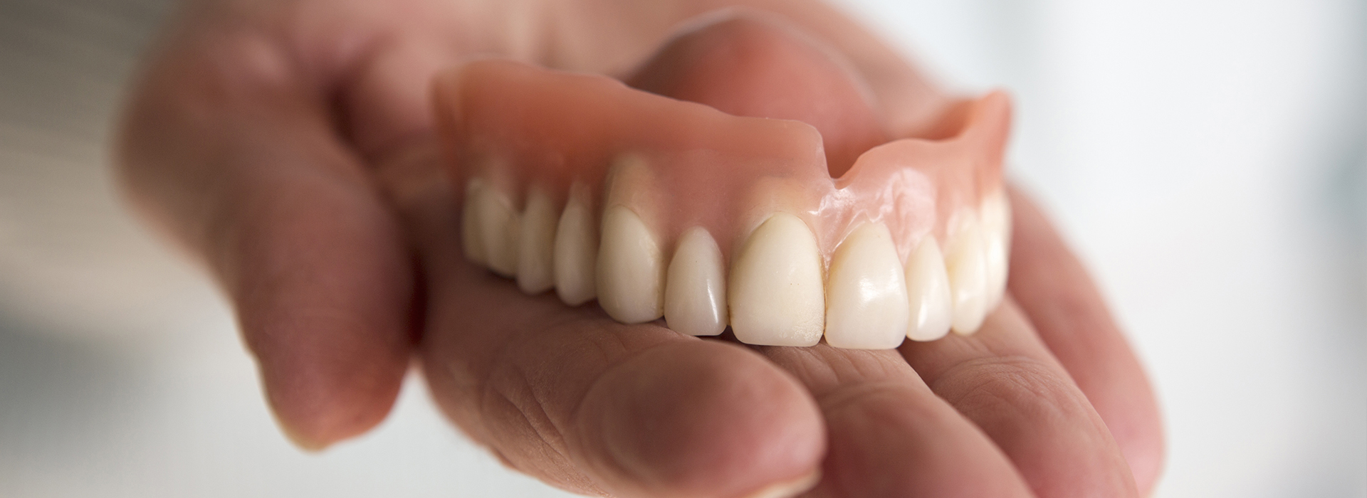 The image shows a person s hand holding a denture plate with several artificial teeth, displayed in front of their face.