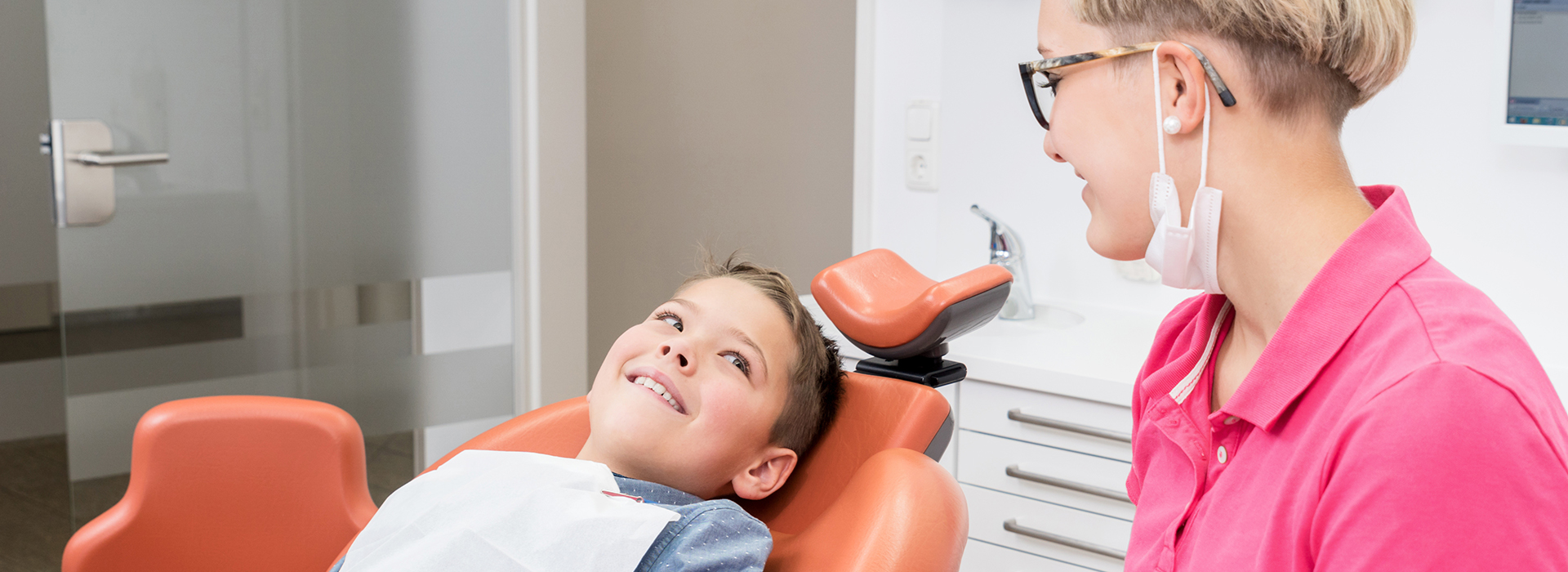 The image shows a dental office scene with a person sitting in the chair, a dentist standing behind them, and a young child seated on the floor smiling.