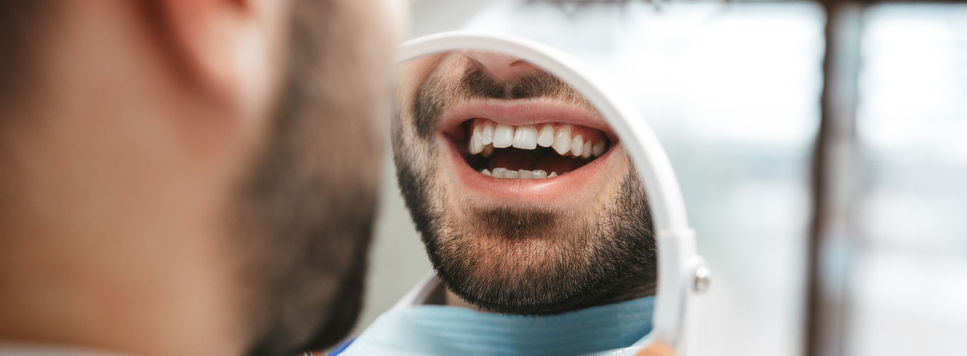 The image shows a man with a beard and mustache, holding what appears to be a small object or tool close to his mouth. He is smiling broadly, revealing teeth, and looking directly at the camera. Another person s face is partially visible on the right side of the photo, with only the nose and lips showing. The background is blurred but suggests an indoor setting with artificial lighting.