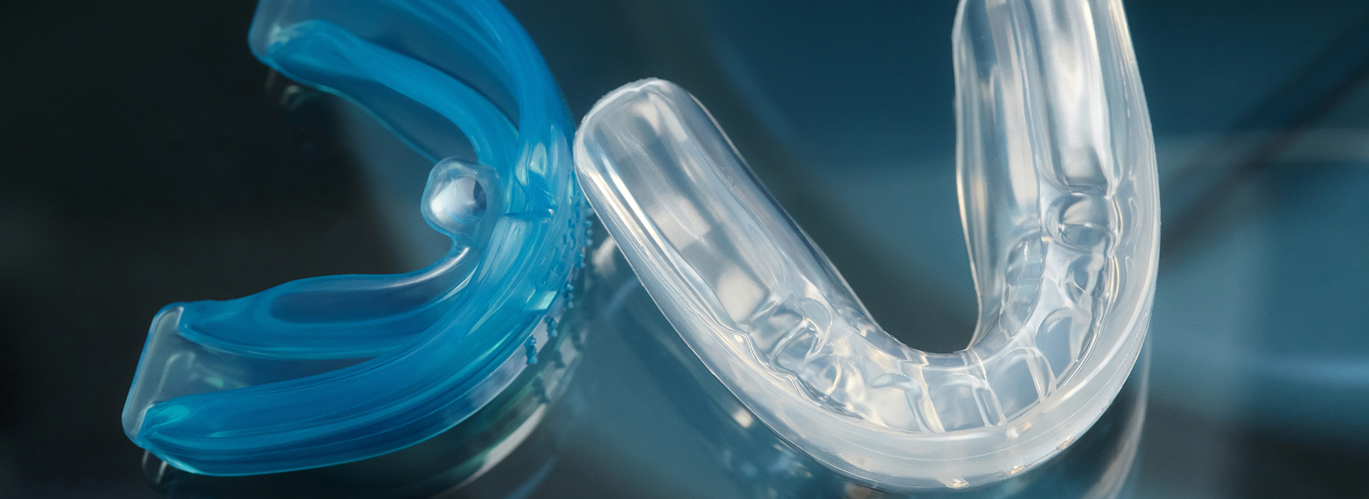 The image shows a close-up of two clear, plastic dental retainers with blue crystals, one partially overlapping the other, placed against a blurred background.