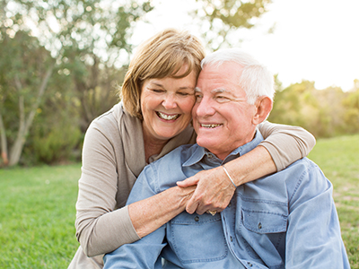 The image shows an older man and woman embracing each other, both smiling, with the man wearing a blue shirt and the woman in a white top. They are outdoors during daylight, possibly on a lawn or park area.