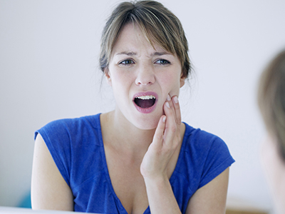 The image is a photograph of a woman with her mouth open, displaying a toothpaste-foamy smile. She appears concerned or alarmed, and there s a reflection of her face in the mirror behind her.