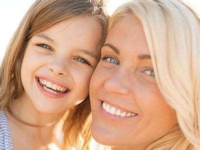 The image depicts a woman and a child, likely a mother and daughter, smiling at the camera with a blurred background.