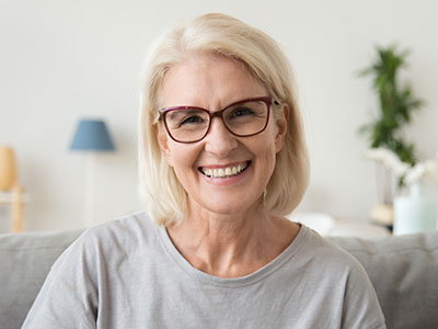 A middle-aged woman with blonde hair, wearing glasses and a gray top, smiling at the camera.