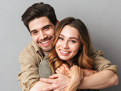 A young couple embracing and smiling, with the man wearing a beige shirt and the woman in a dark top.