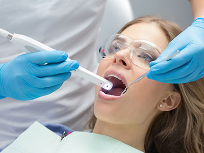 A dental professional using a device to clean or examine a patient s teeth, with the patient seated in a dental chair and wearing blue gloves.