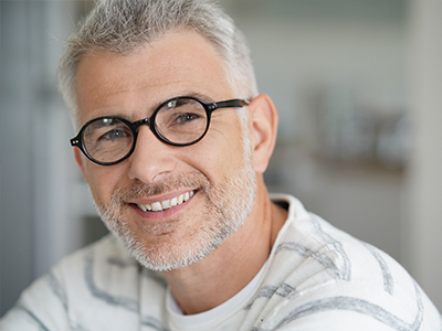 The image shows a middle-aged man with glasses, smiling and looking directly at the camera. He has short gray hair and is wearing a white shirt with horizontal stripes.