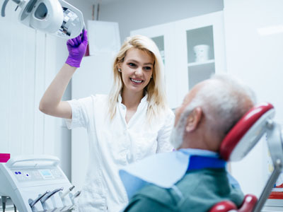 A woman in a white coat is standing next to an older man seated in a dental chair, holding his chin as she appears to be examining him.