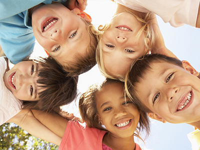 The image features a group of children, predominantly smiling and looking towards the camera, with one child appearing to be leaning forward. They are outdoors under clear skies, wearing casual clothing suitable for warm weather.