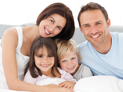 A family of four, including two adults and two children, smiling in a cozy bedroom setting with one adult lying down and the other sitting up.