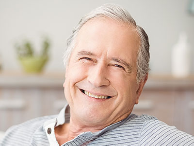 The image depicts a smiling older man with gray hair, wearing glasses and a blue shirt, sitting in a chair indoors.