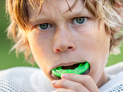 The image features a young individual with blonde hair, wearing a sports jersey and holding a green object in their mouth.