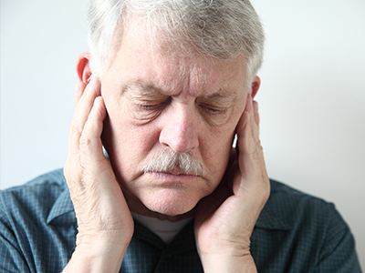 The image is a photograph of an older man with his hand on his face, appearing to be in pain or discomfort. He has grey hair and is wearing a dark shirt. There is no visible text within the image.